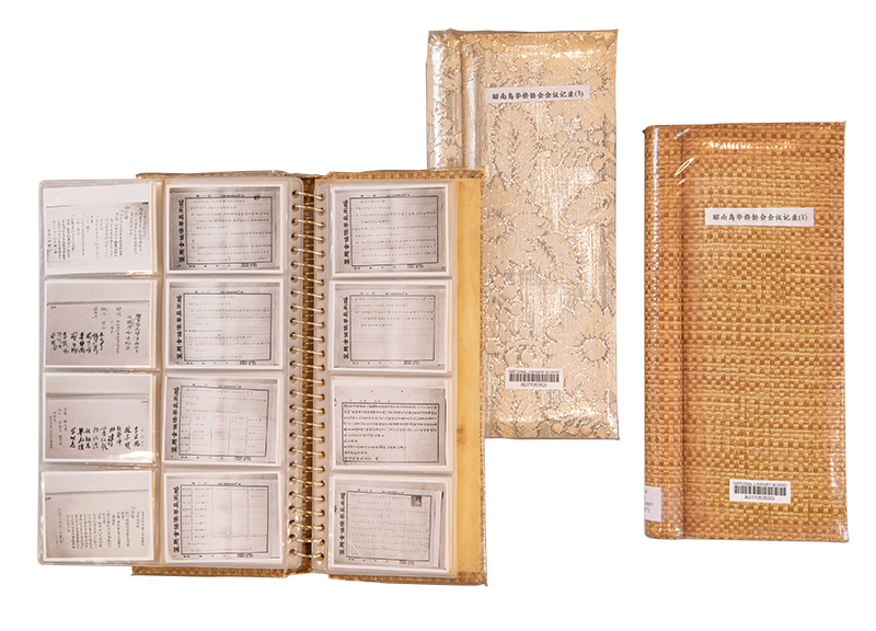 An open binder with handwritten pages and two fabric-covered books placed alongside.